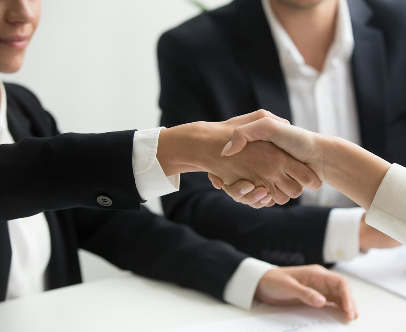 Close-up of two people shaking hands over a white table, with a third person in a suit visible in the background.