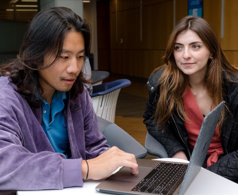 Two students, one in a purple velvet jacket and the other in a black quilted coat, looking at a laptop screen together indoors.