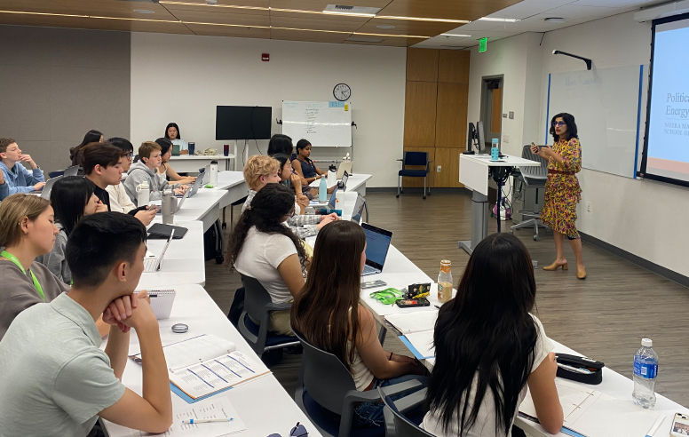 A female instructor in a yellow floral dress stands at the front of a classroom lecturing to a group of students seated at long tables with laptops.