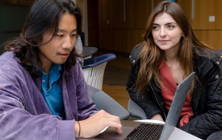 Two students, one in a purple velvet jacket and the other in a black quilted coat, looking at a laptop screen together indoors.