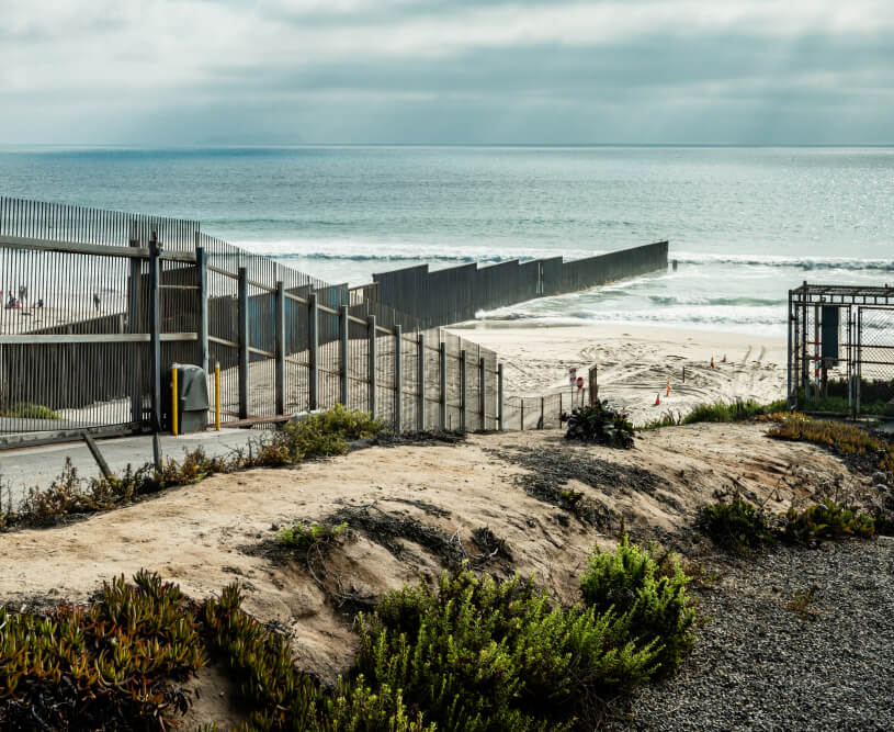 View of the U.S.-Mexico border fence extending into the Pacific Ocean, with a sandy beach and coastal plants in the foreground.