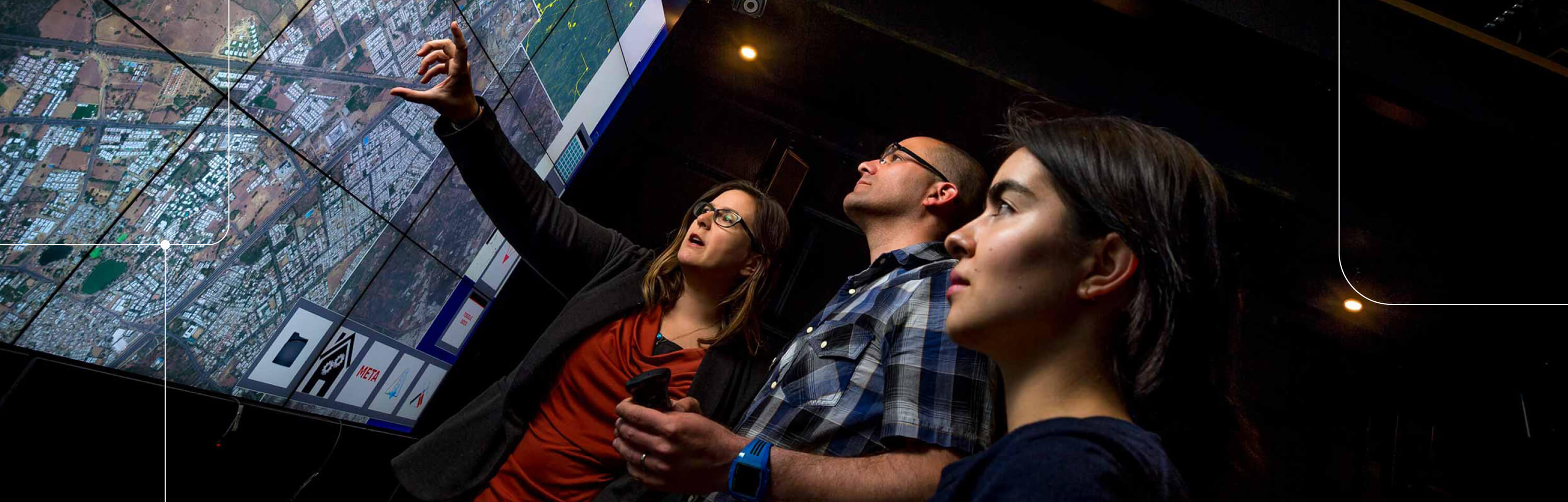 Three people in front of numerous screens showing a visualization of geographic information system data
