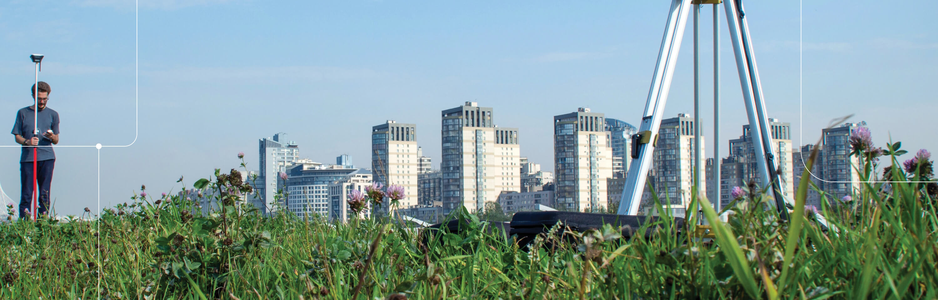 City skyline with grass and a land survey tripod in the foreground, while a land surveyor is taking measurements